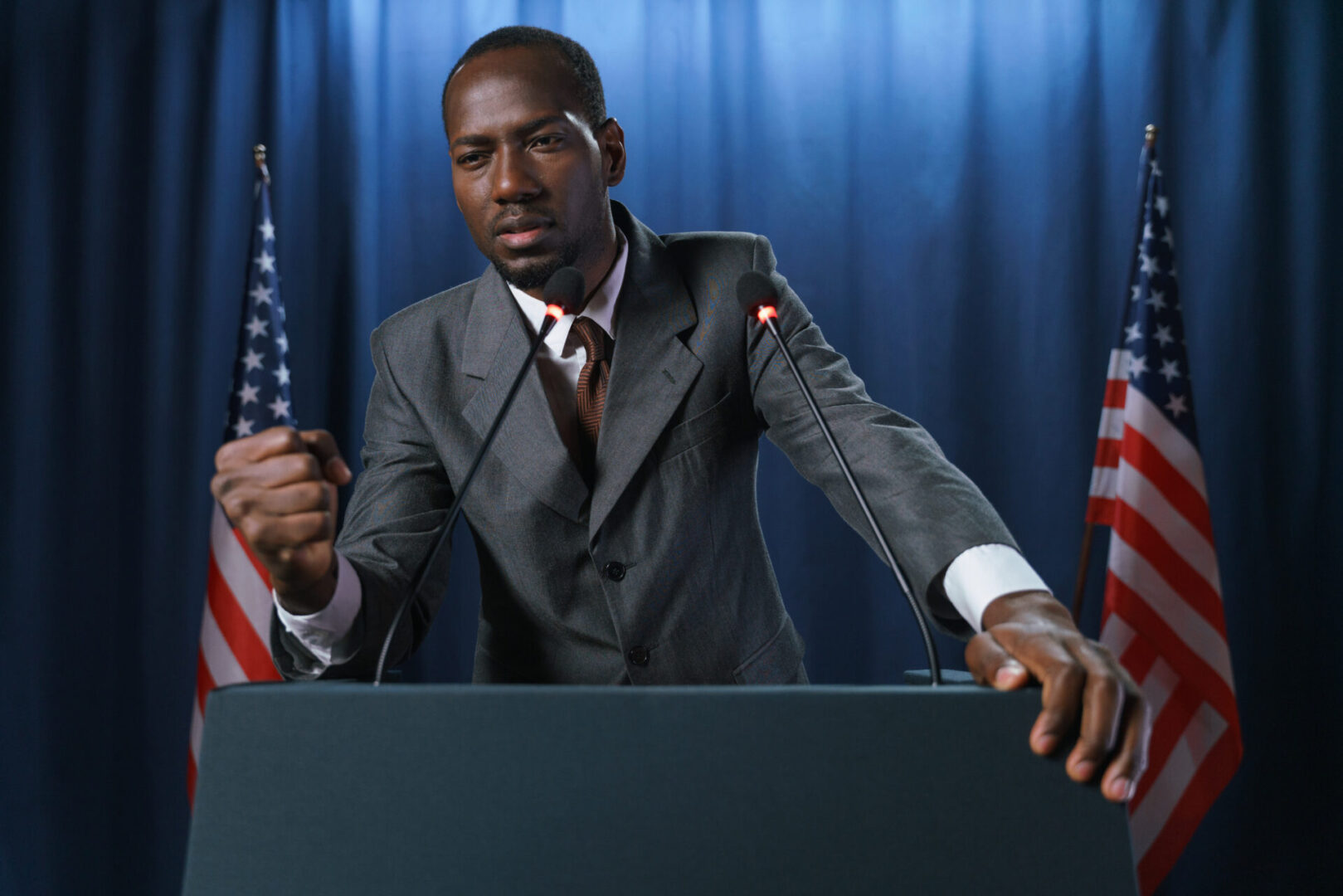Serious African-American politician in a gray suit gives a speech, gesturing with a fist, standing against the blue background with American flags, we see him from the waist up