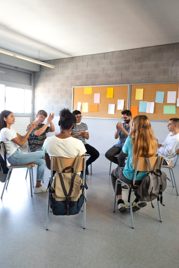 Multiracial group of high school students sitting in a circle clapping together celebrating achievement. Support group. Vertical image. Mental health concept.