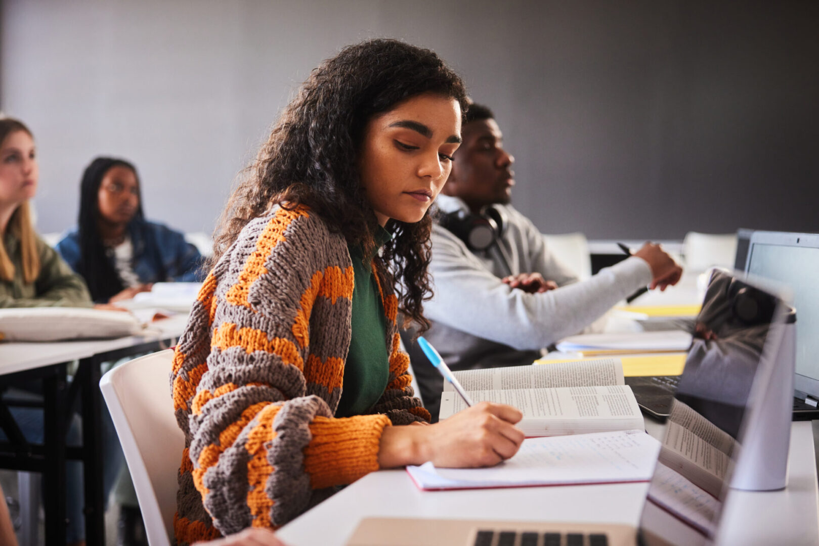 Focused young female college student writing down notes while sitting at a table during a lecture with classmates at school