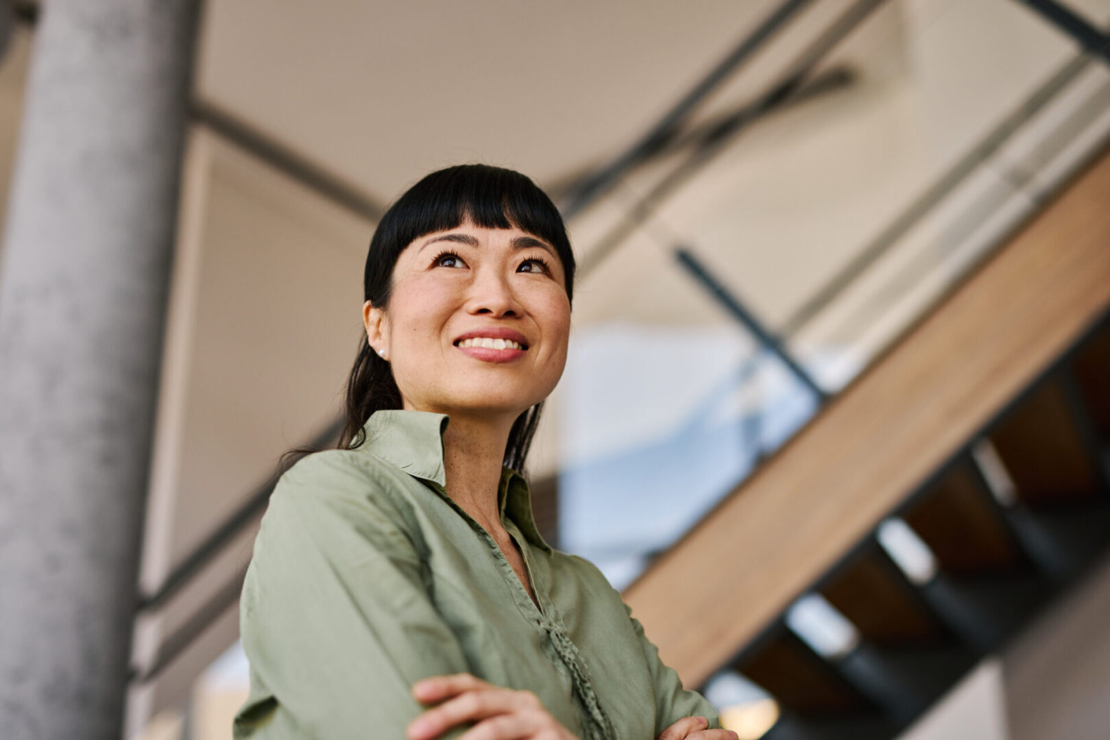 Low angle view of a smiling mid-adult Asian businesswoman with arms crossed, gazing upward in a modern office building, exuding confidence and ambition while contemplating future opportunities