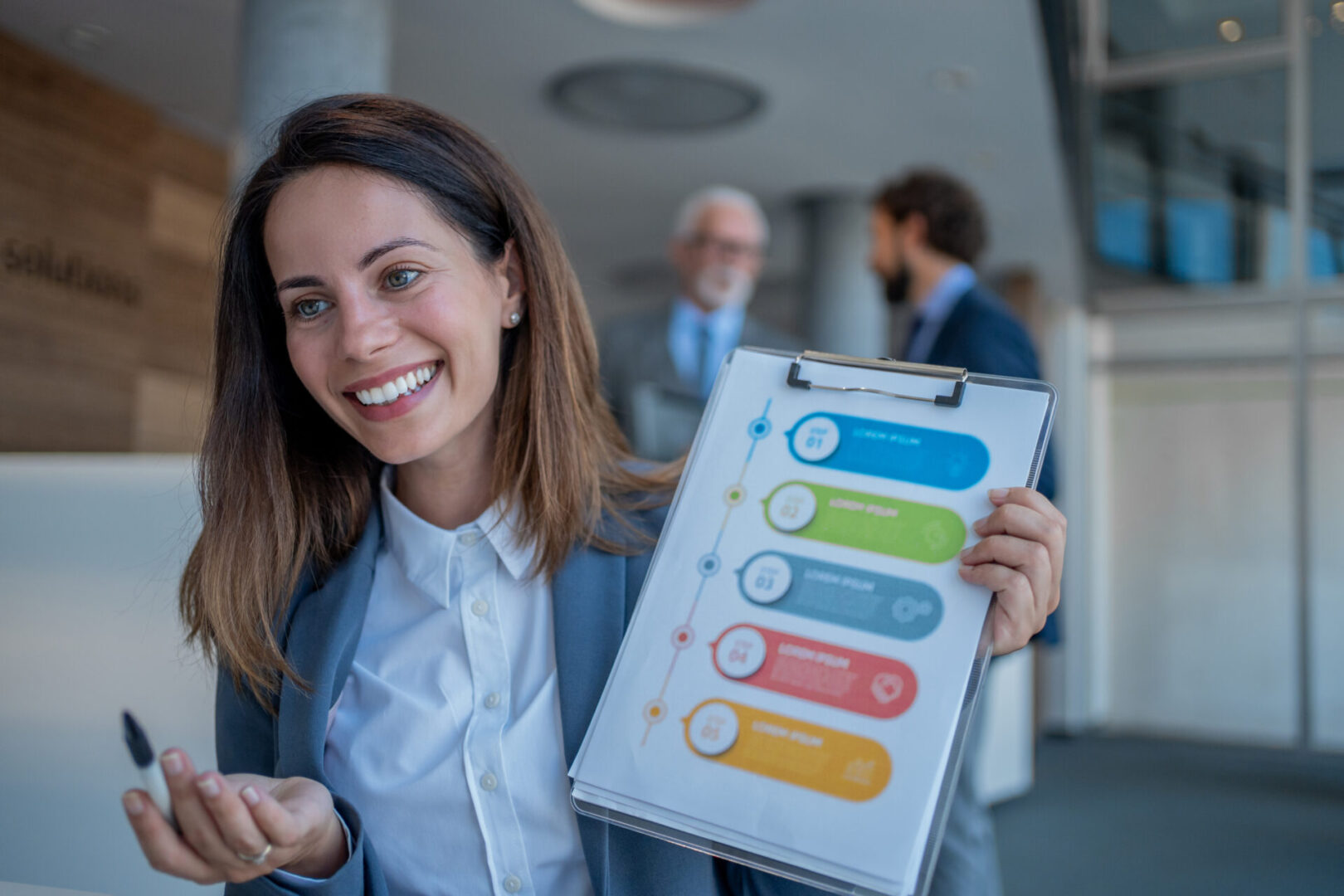 Young businesswoman holding a clipboard and showcasing infographic steps, smiling while explaining strategy to colleagues in a collaborative office environment