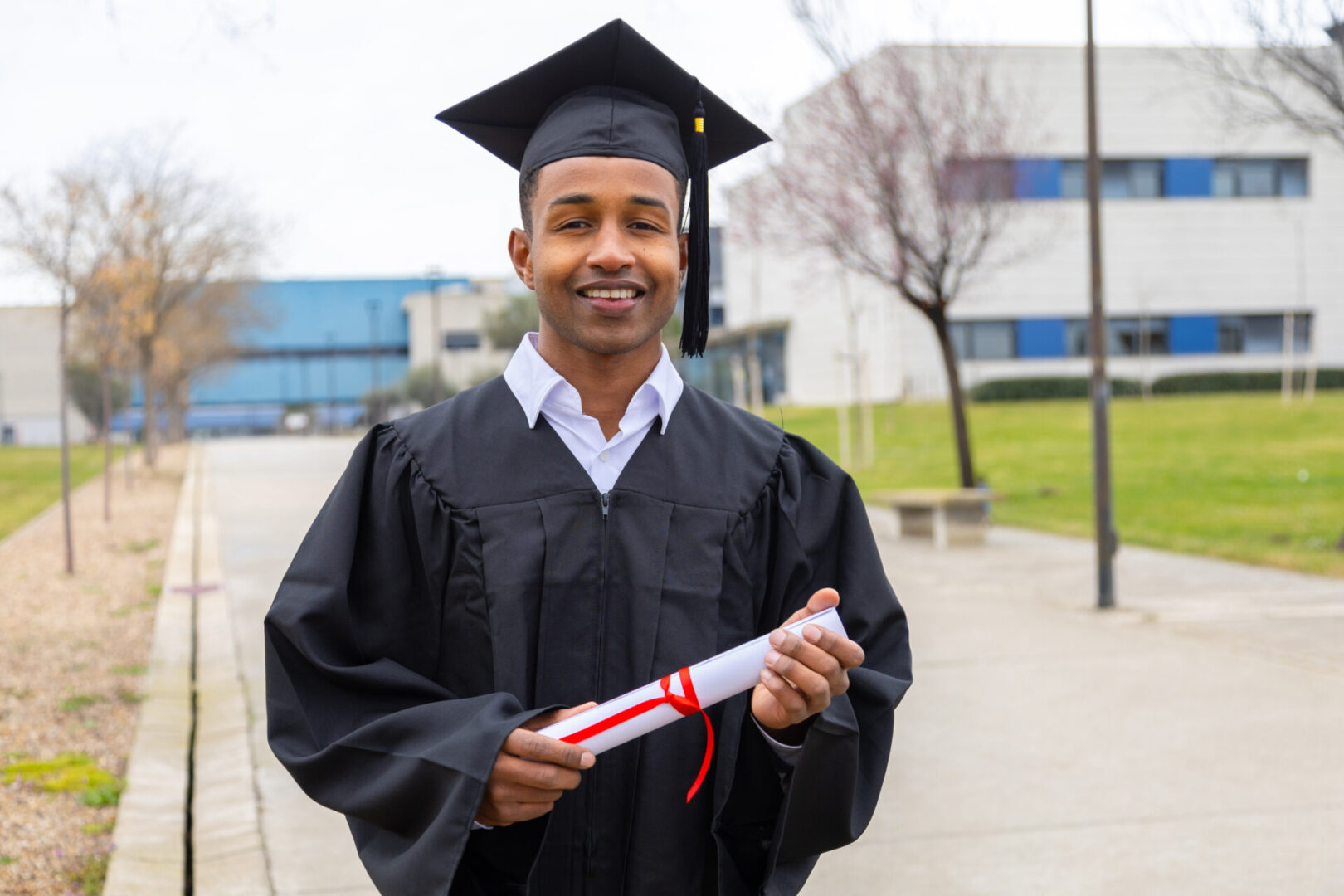 Smiling african american graduate student wearing graduation gown and cap holding diploma walking in university campus