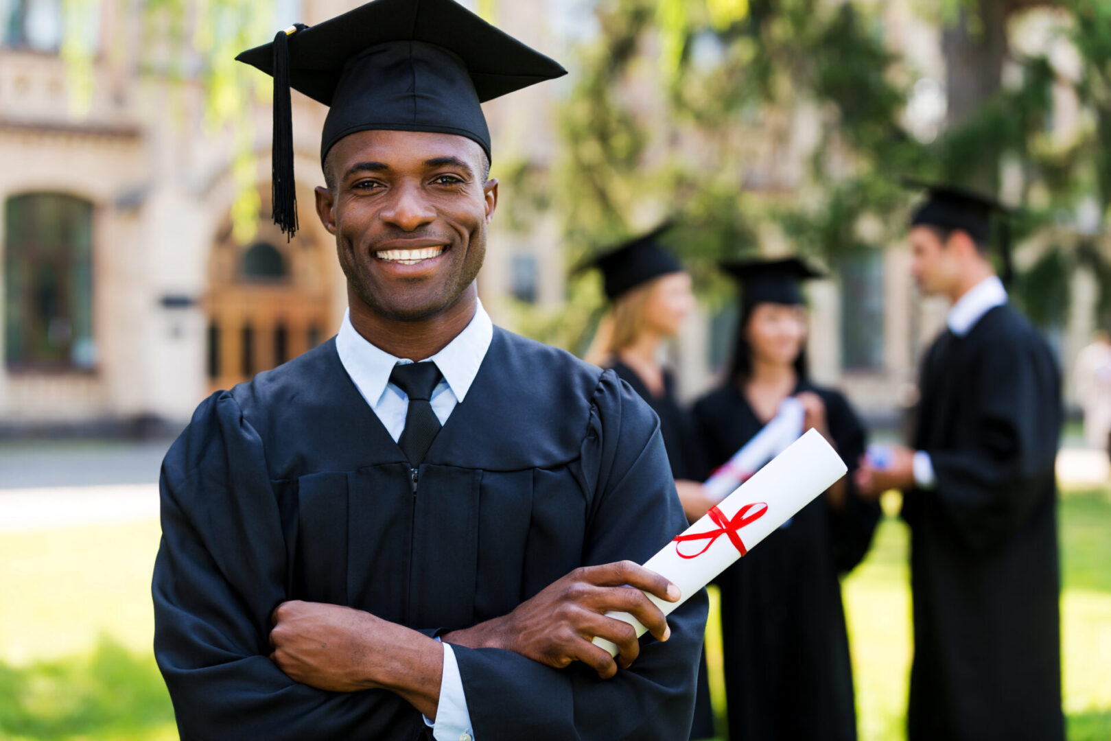 Happy African man in graduation gowns holding diploma and smiling while his friends standing in the background